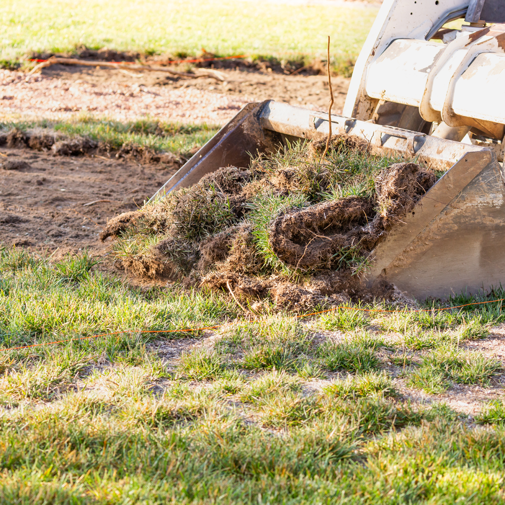 A white skid-steer loader clearing a grass patch to begin digging out a level DIY fire pit area in a backyard.